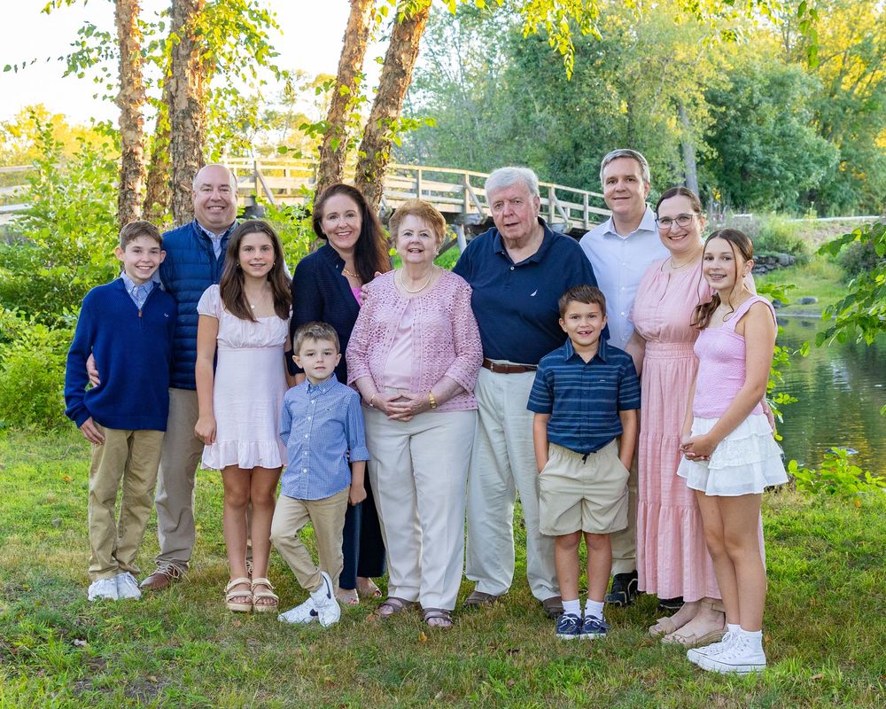 Extended family wearing coordinated blue and pink outfits poses together outdoors in front of trees and North Bridge in Concord, MA.