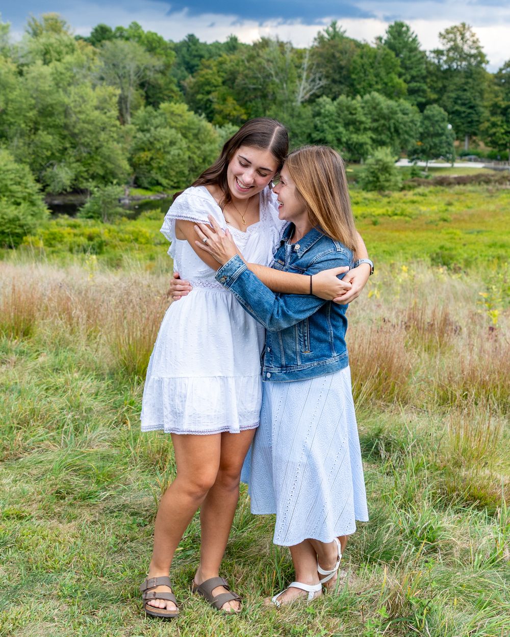 Mother & daughter embrace in a grassy meadow on a sunny summer day, wearing white and denim outfits during high school senior portraits.