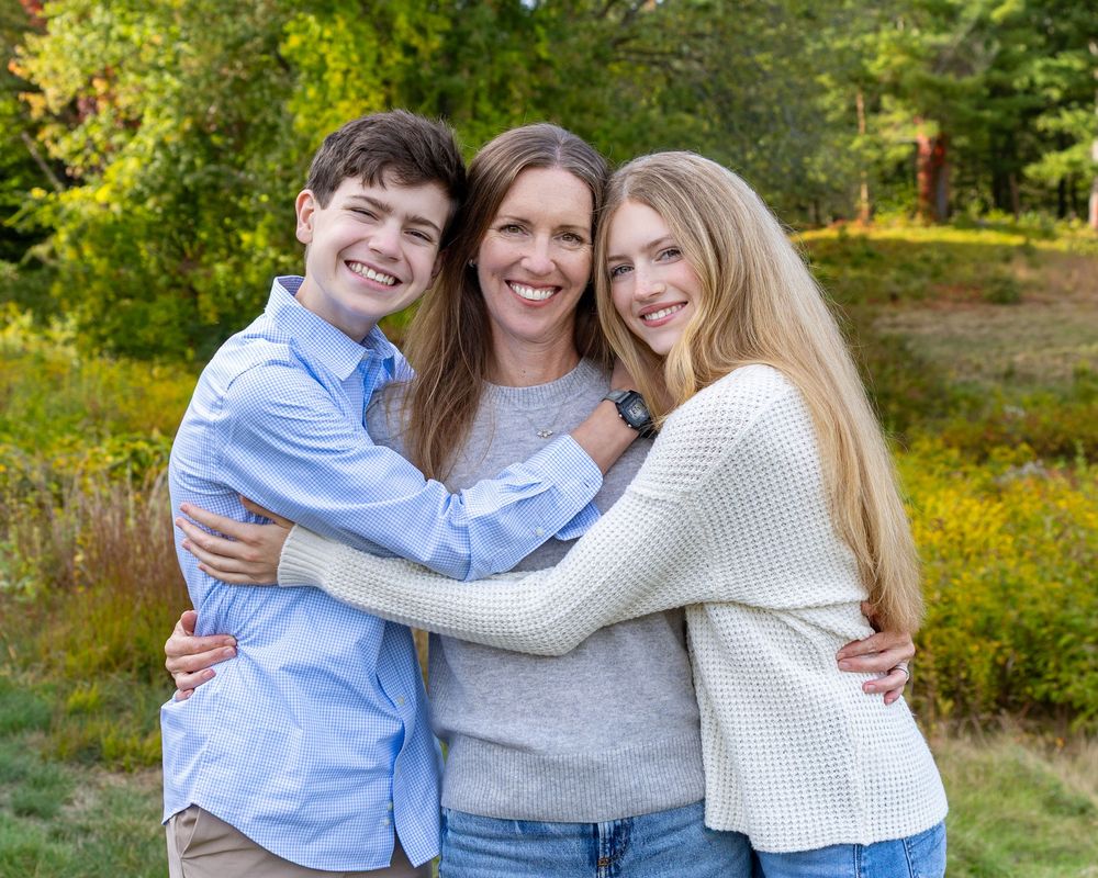 Mom & kids in light blue and white sweaters embraces while posing outdoors among autumn foliage & greenery.