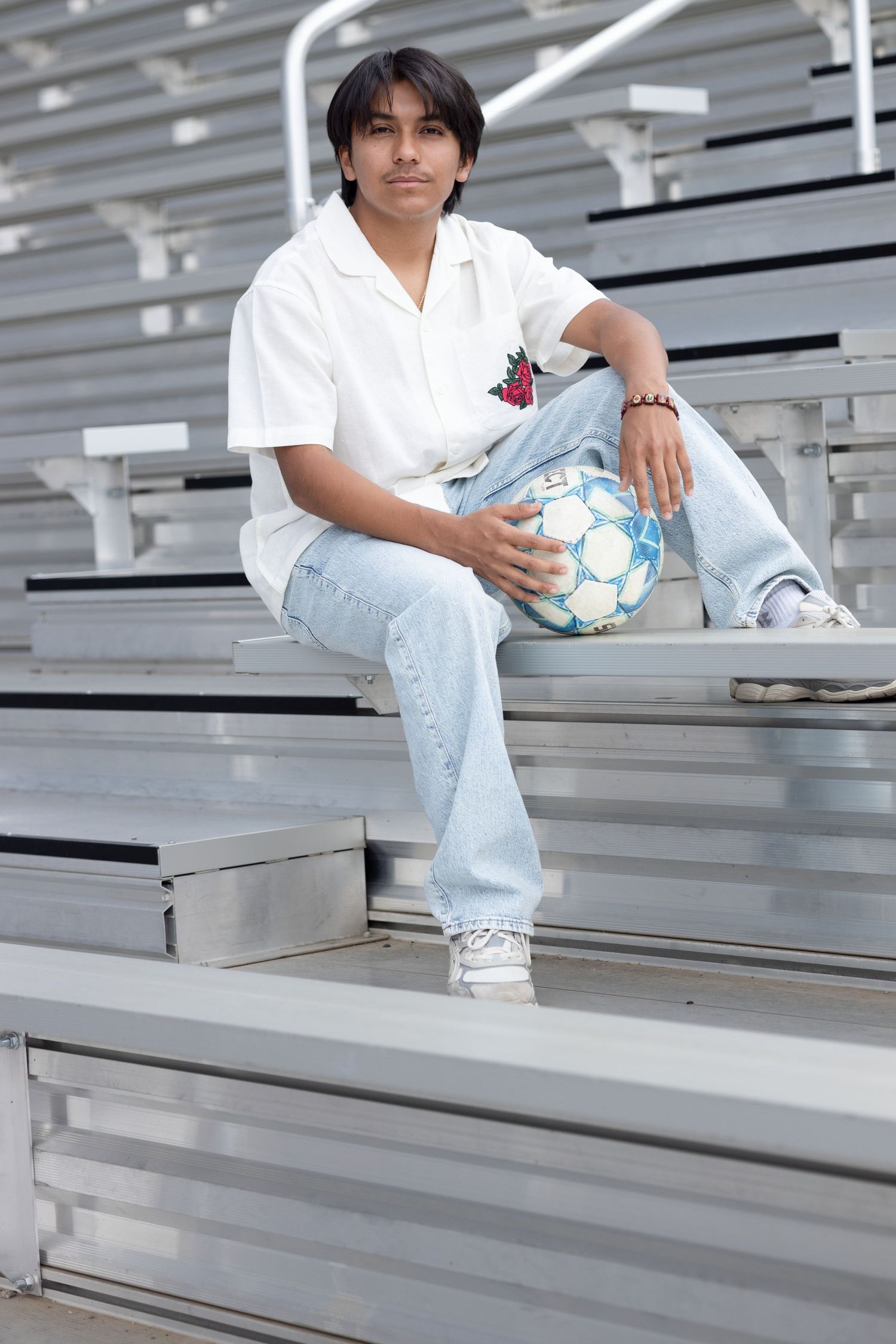 A student athlete in a white shirt and light jeans sits casually on bleachers holding a soccer ball.