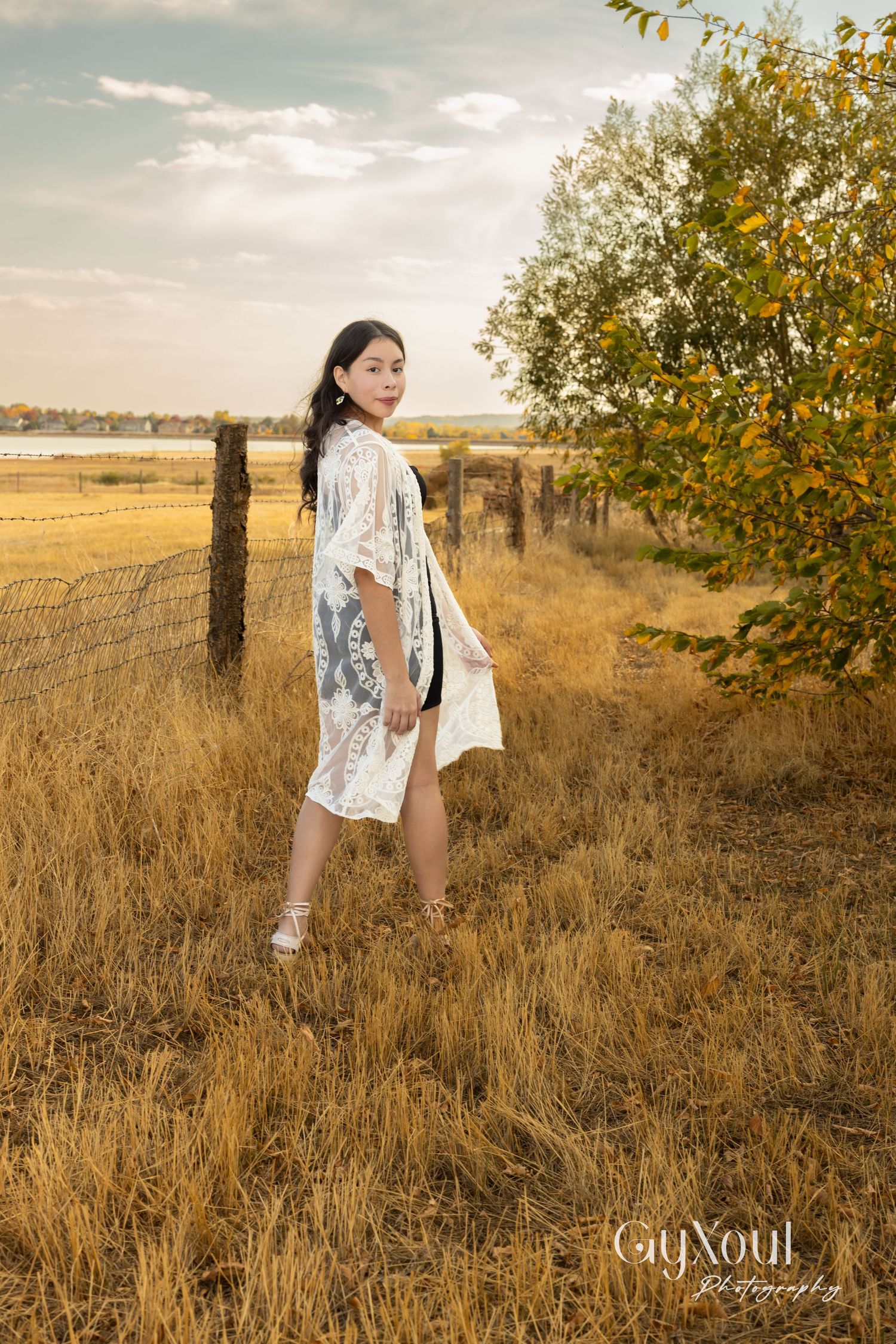 Figure in white lace kimono poses in golden field at sunset with rustic fence and autumn trees.