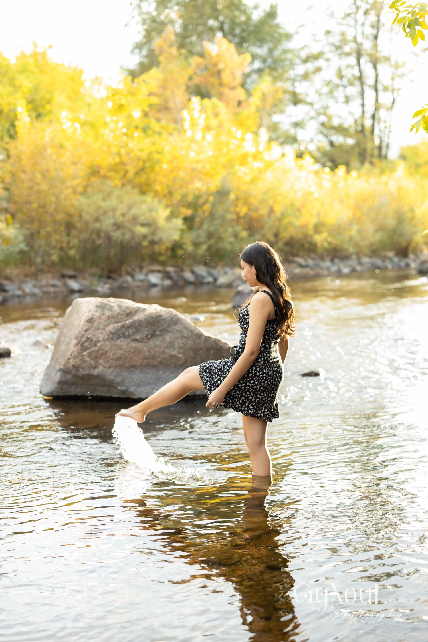 Person in polka dot dress splashing in river water during golden hour near large boulder.