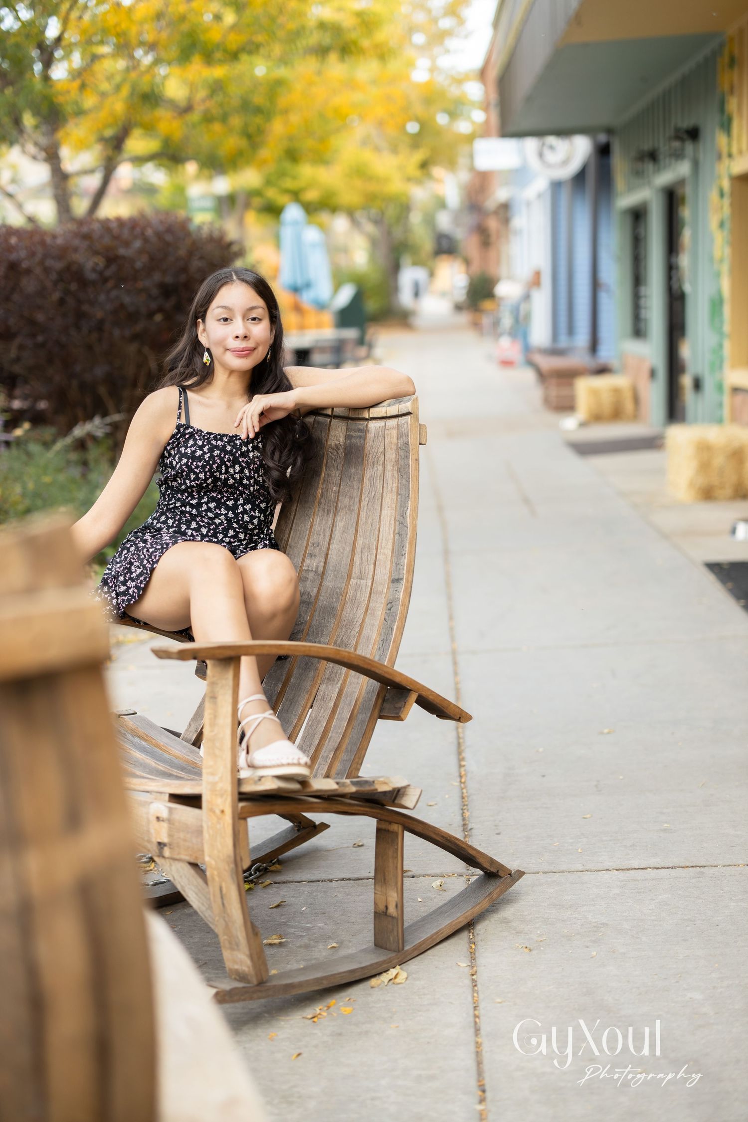 Person relaxing in wooden rocking chair on charming small-town sidewalk with autumn trees.