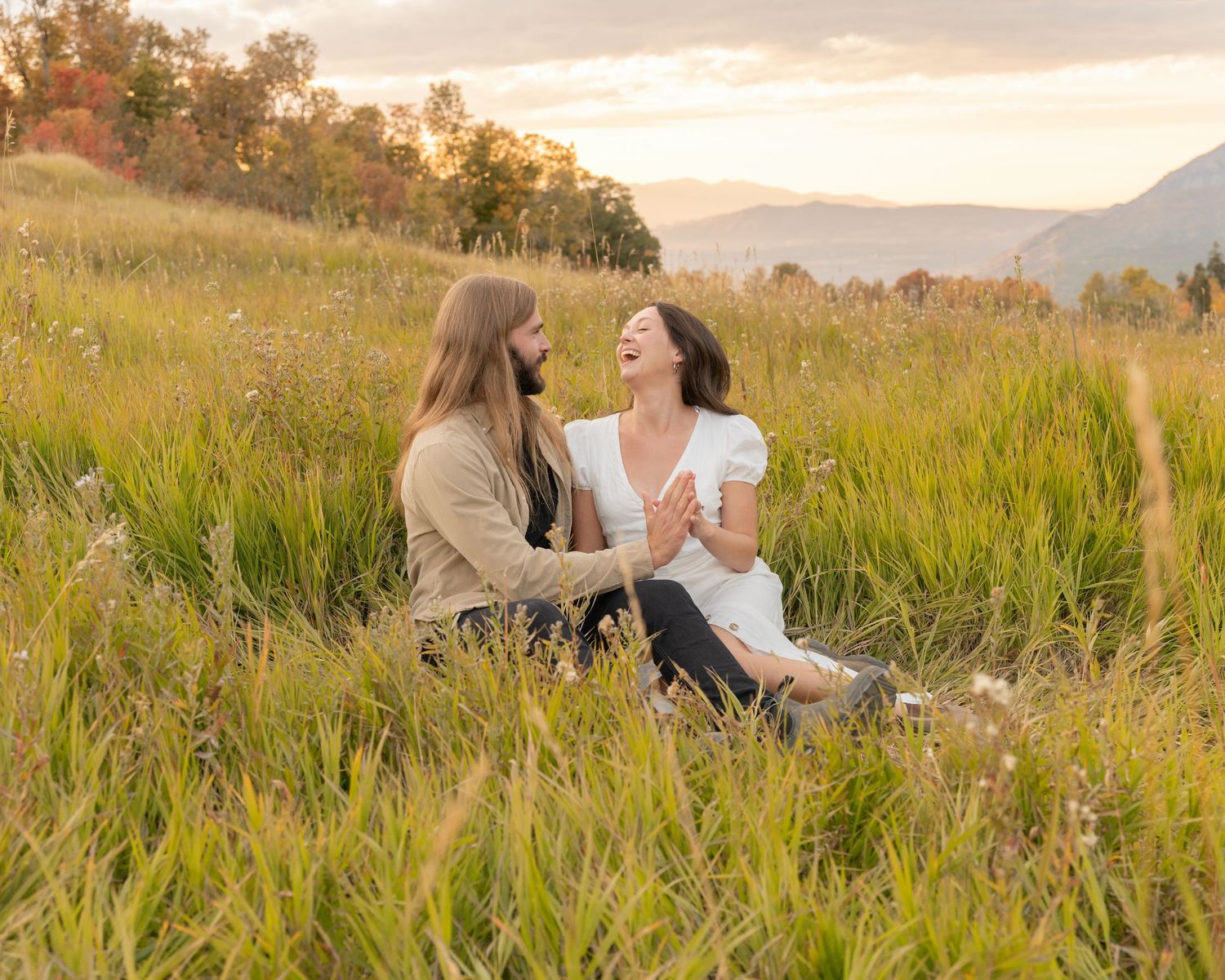 Two people sit together in a golden field at sunset sharing intimate moments among tall grass and mountains.
