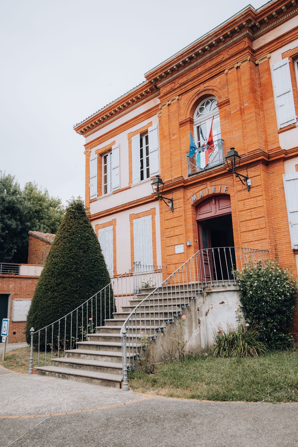 Historic orange brick French mairie building with stairs, flags, and trimmed hedges.