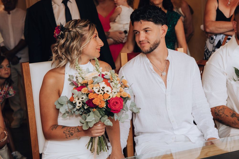 Smiling bride holding colorful bouquet sitting beside groom in white shirt during wedding ceremony.