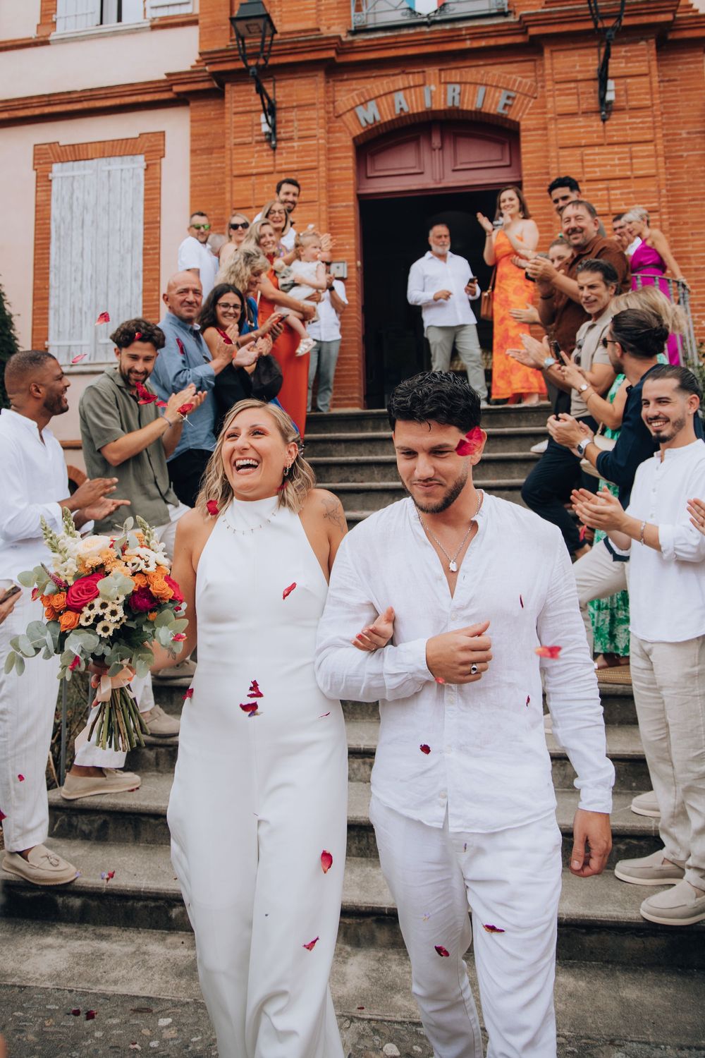 Joyful newlyweds walking down steps of a mairie as guests shower them with red petals.