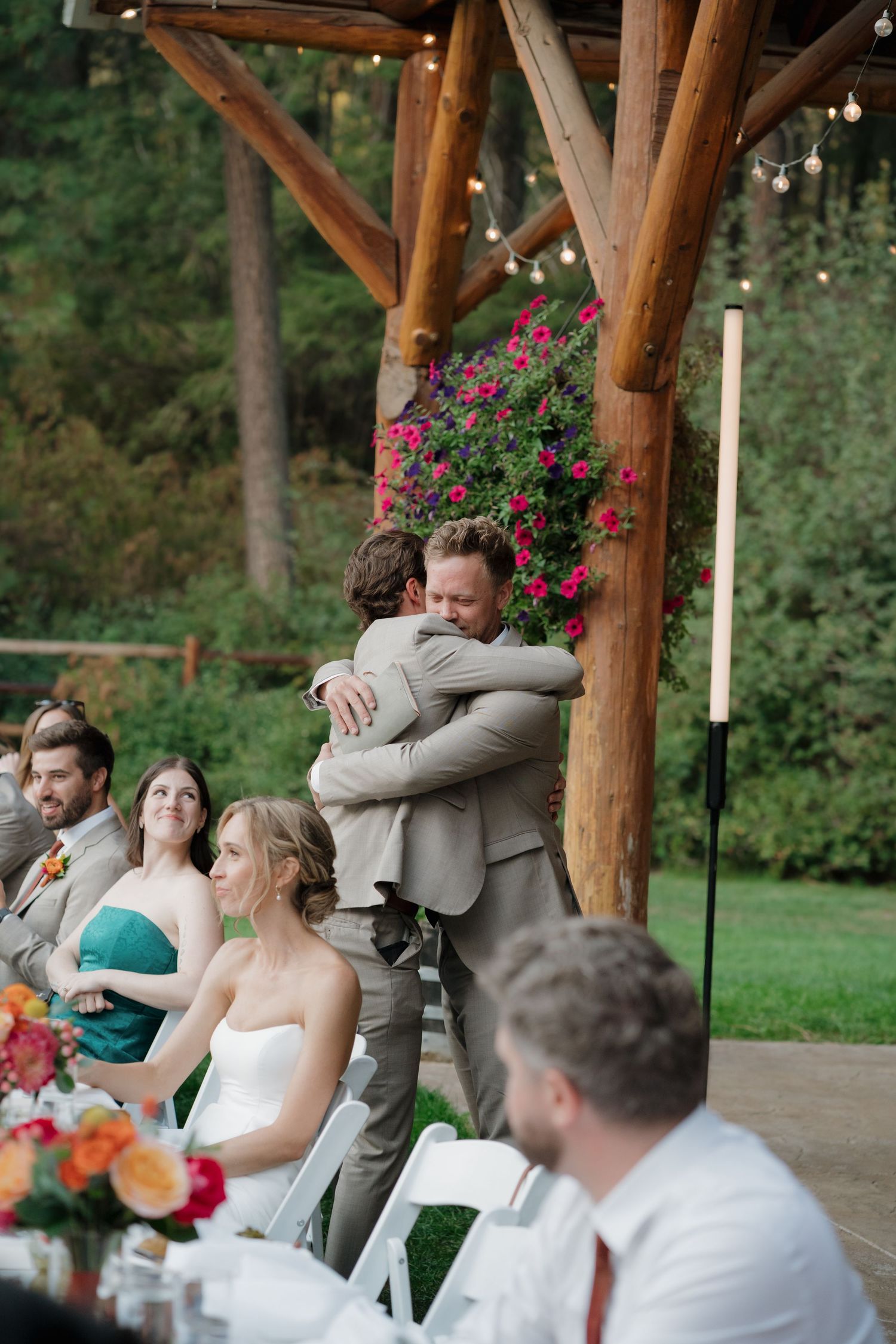 Wedding guests gather under a rustic wooden pavilion decorated with hanging flower baskets during an outdoor reception.
