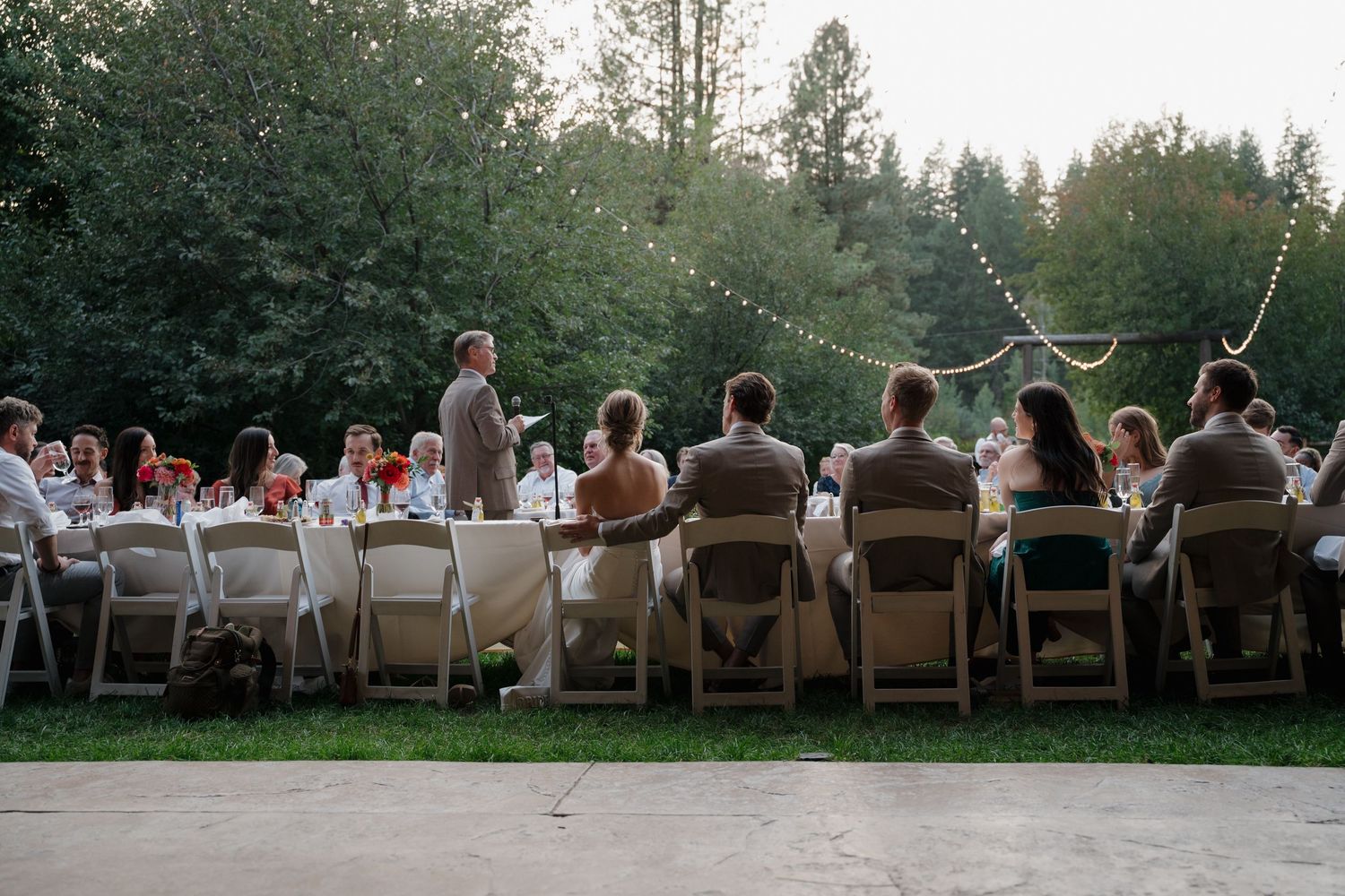A group of wedding guests sitting in wooden chairs facing a scenic forest view during an outdoor ceremony.