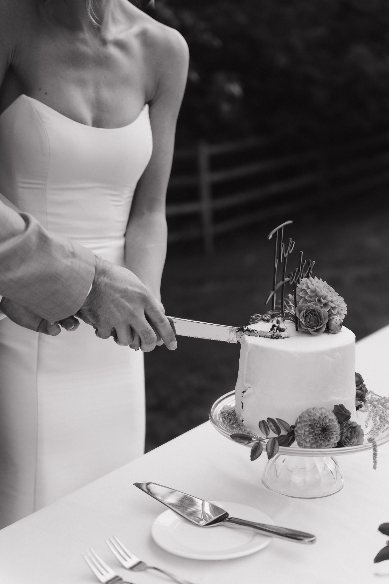Black and white photo of someone in a white dress cutting into an elegant tiered wedding cake decorated with flowers.