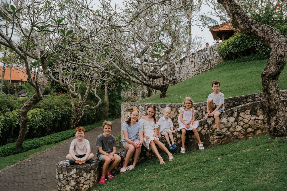 A group sits together on a stone wall under gnarled trees on a lush hillside garden setting.