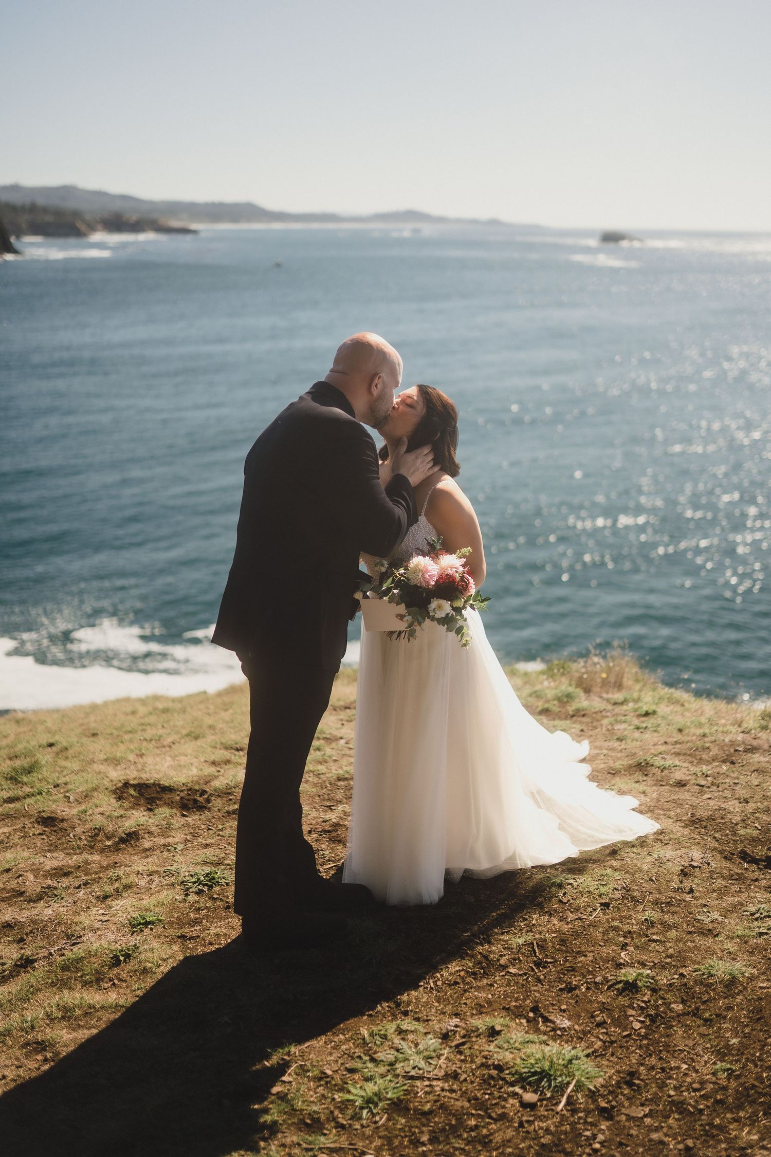 A couple shares an intimate moment on a scenic coastal cliff overlooking sparkling blue ocean waters.