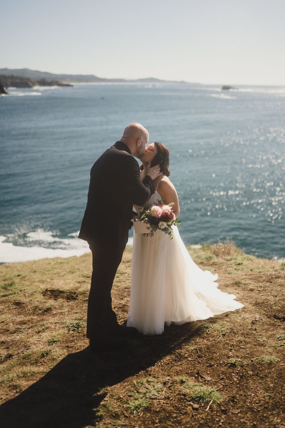 A couple shares an intimate moment on a scenic coastal cliff overlooking sparkling blue ocean waters.