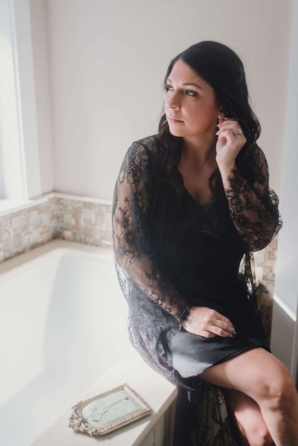 Woman in black lace dress sits thoughtfully by bright window in elegant boudoir photo series.
