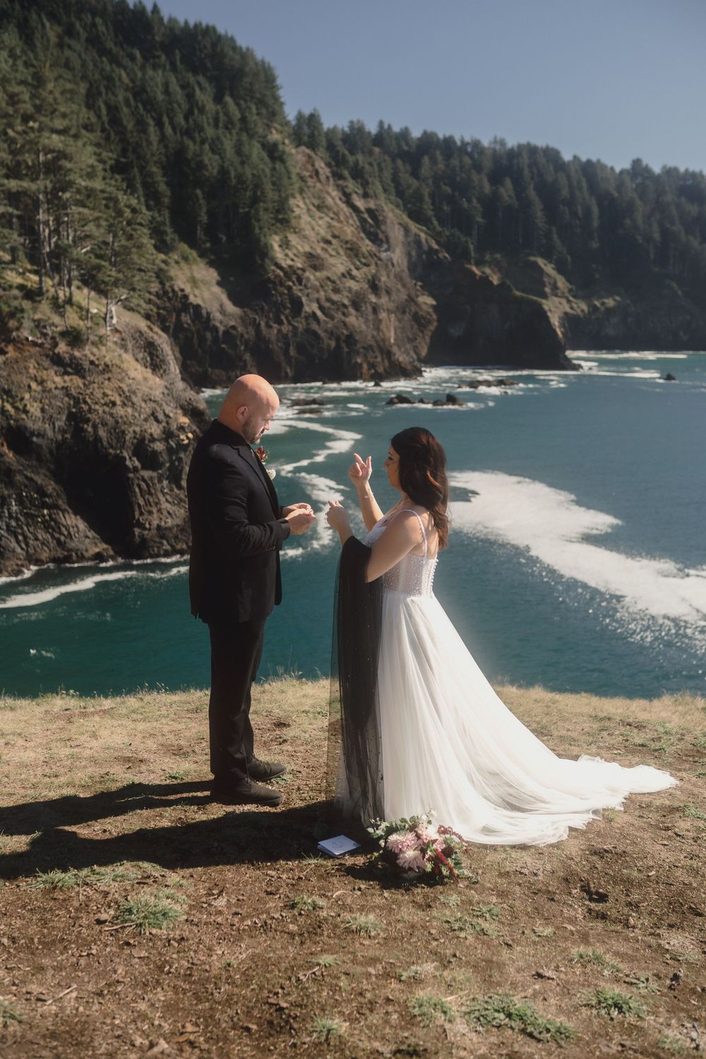 A couple stands at the edge of a dramatic coastal cliff overlooking the Pacific Ocean during an intimate wedding ceremony.