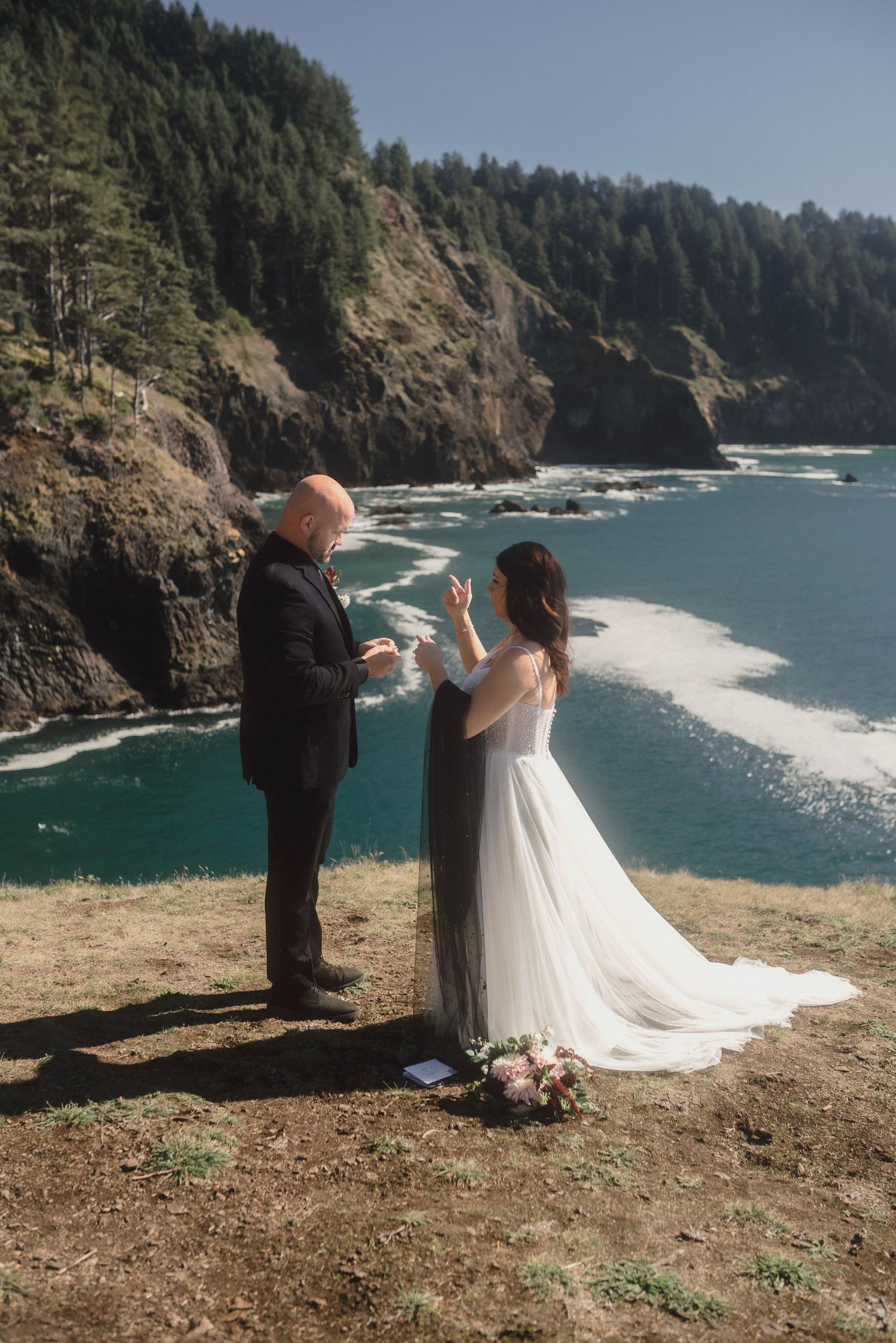 A couple stands at the edge of a dramatic coastal cliff overlooking the Pacific Ocean during an intimate wedding ceremony.