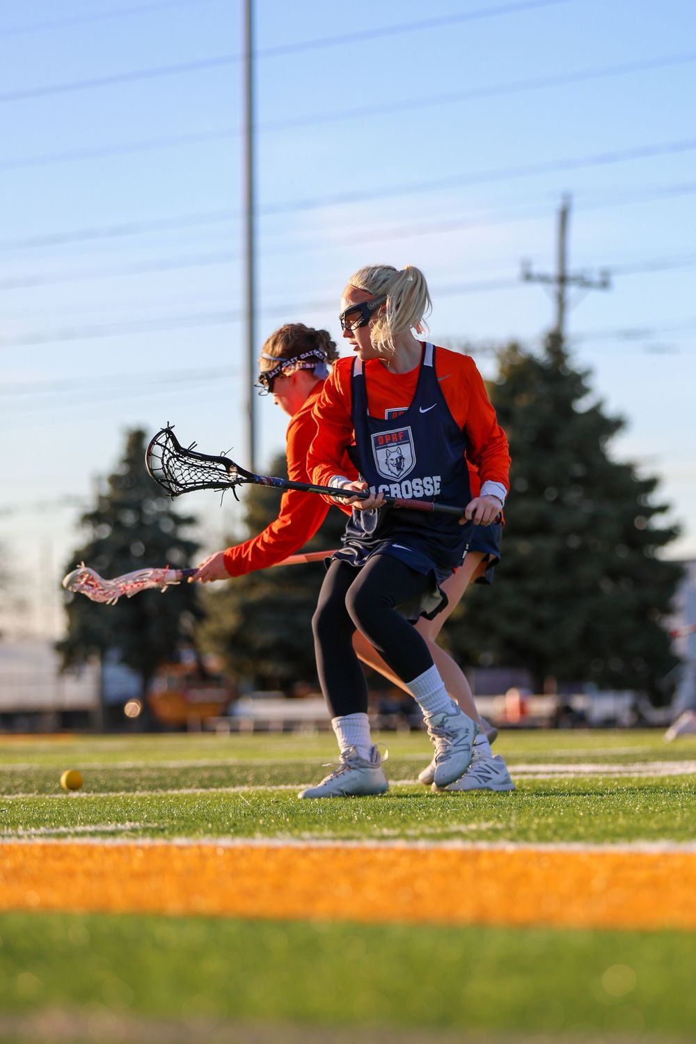 Lacrosse players in orange jerseys practice ground ball drills on a sunny field.