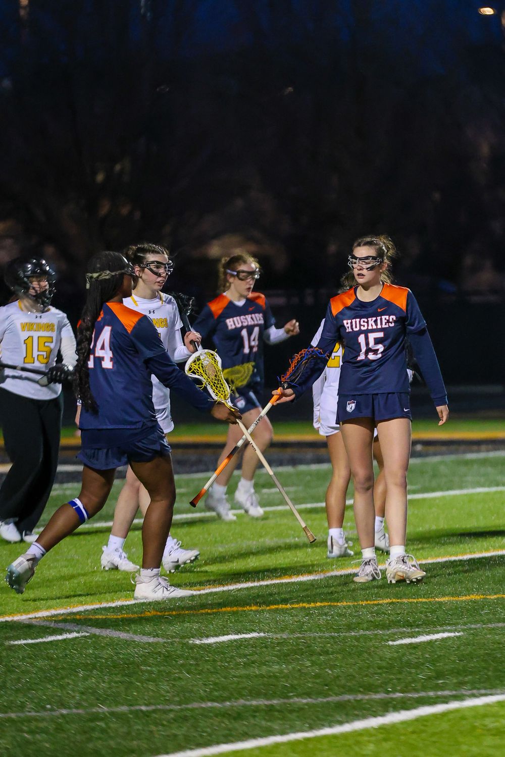 Women's lacrosse team players stretch on a field at night during practice.