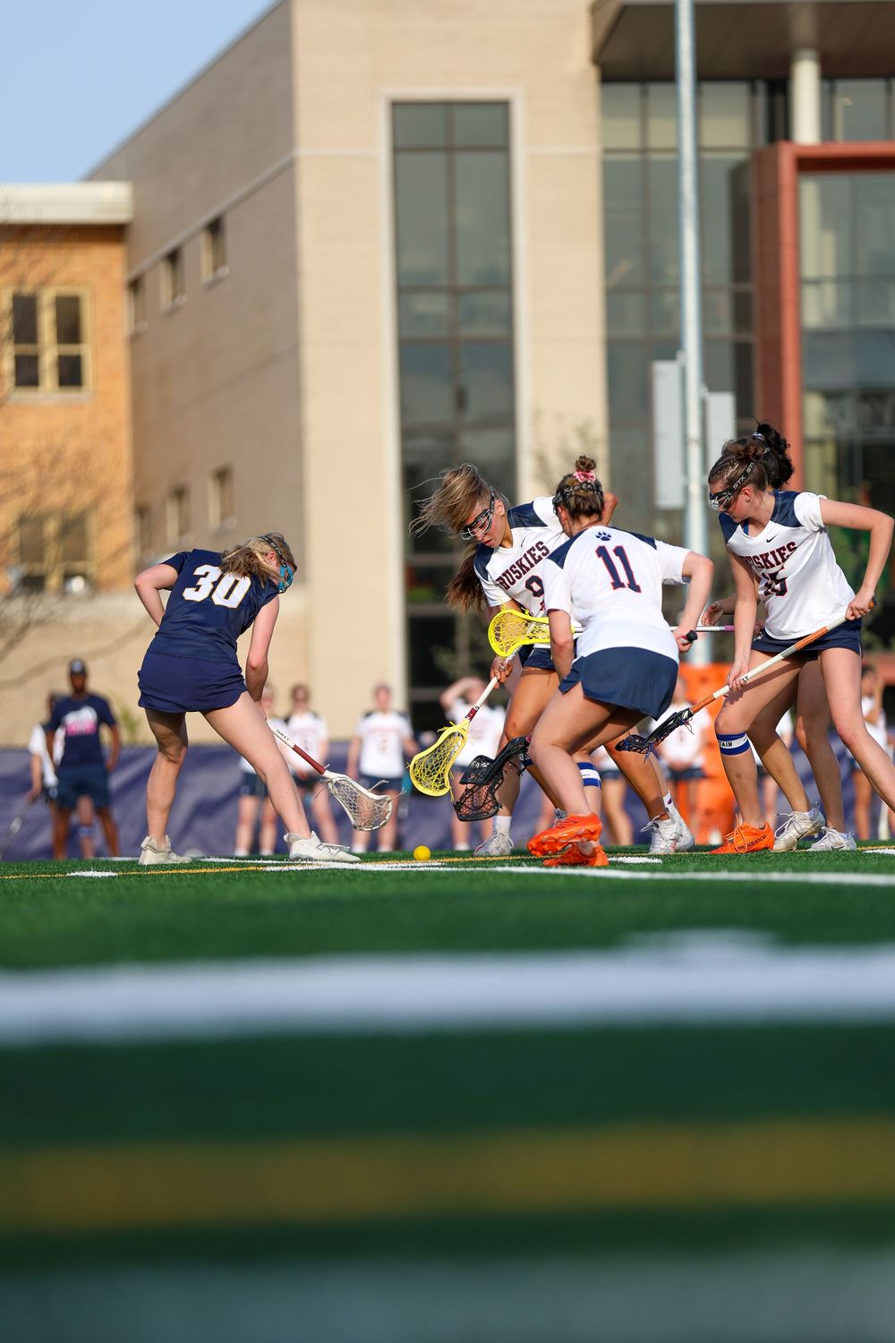 Athletes doing warmup exercises on turf field with campus buildings in background.