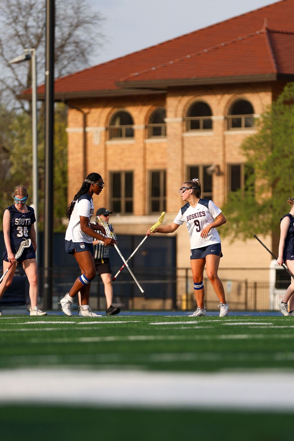 Lacrosse players perform dynamic stretches on a field in front of brick buildings.