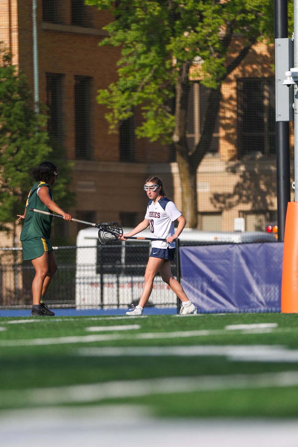 Athletes practicing lacrosse drills on turf field near campus buildings.