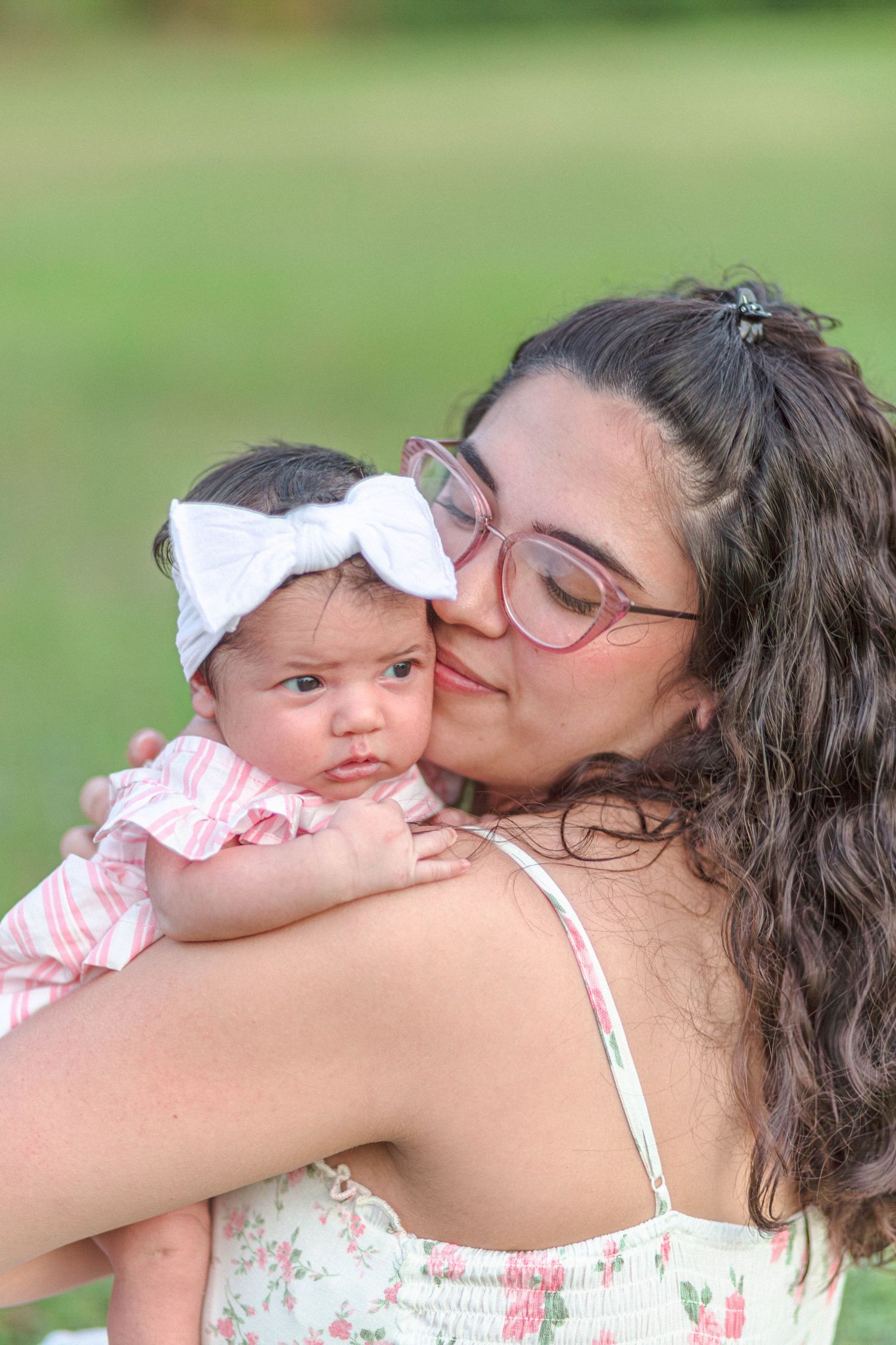 A young mother with glasses kisses her newborn baby girl wearing a white bow headband outdoors.