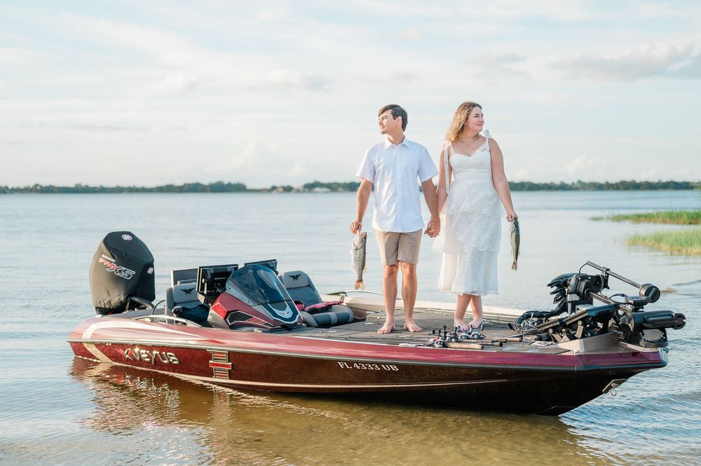 A couple stands together on a red bass fishing boat on calm waters during a bright summer day.