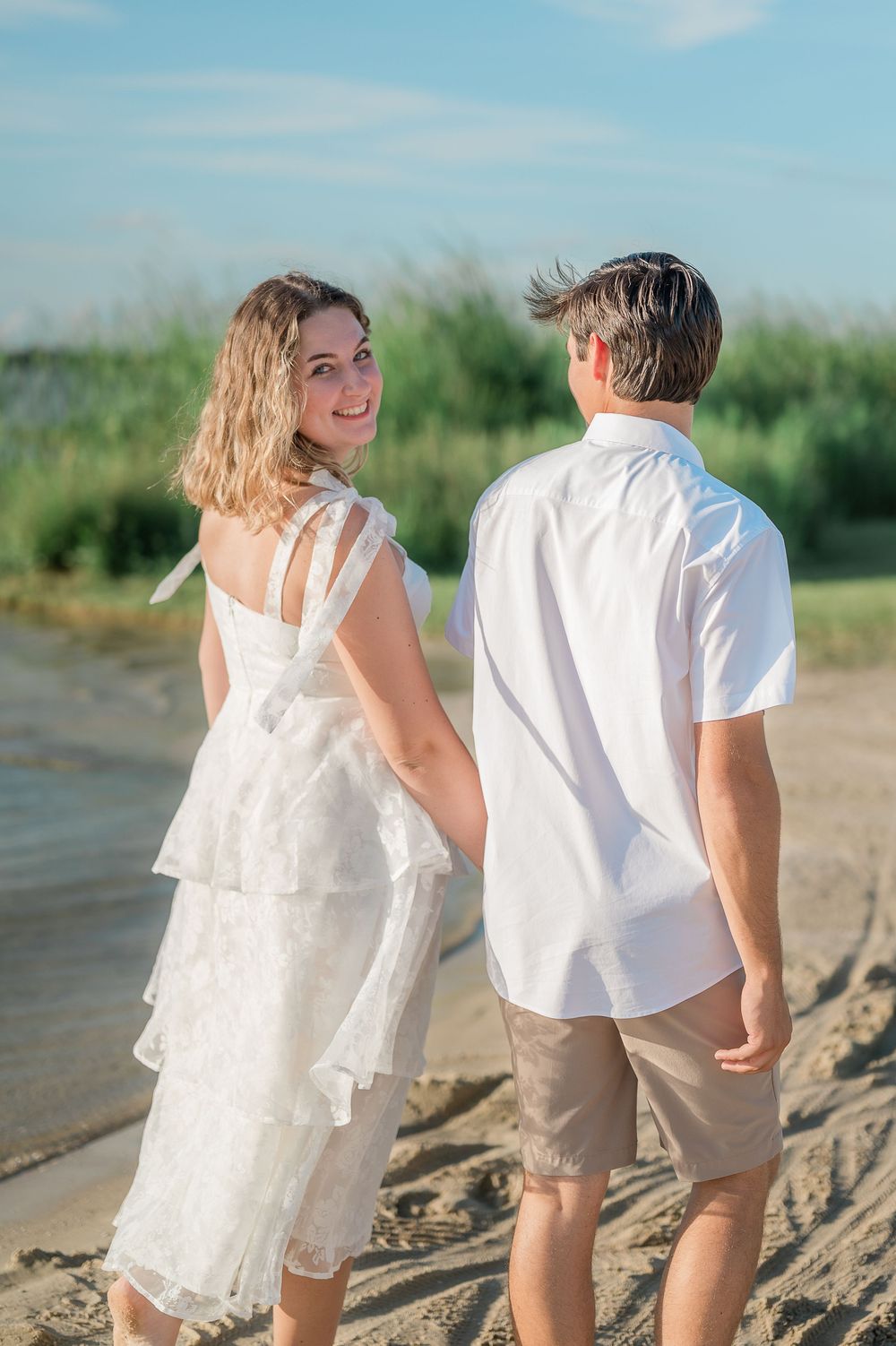 A couple in white outfits stands together on a sandy beach with water and greenery in the background.