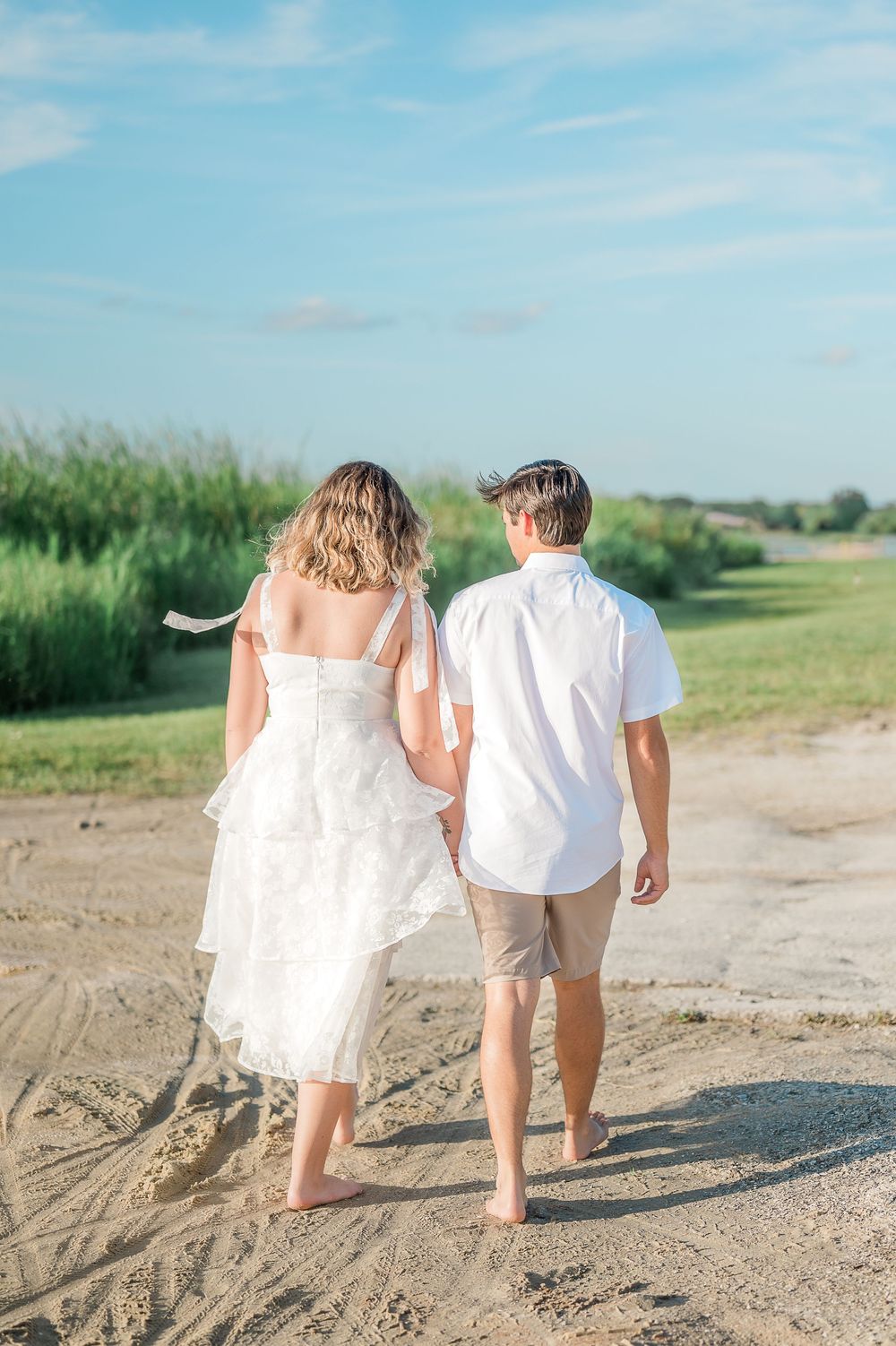 A couple walks barefoot on a sandy beach path with green grass and blue sky on a sunny summer day.
