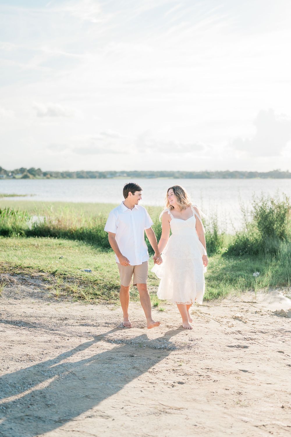 A couple walks together on a sandy beach path during a bright summer day, creating long shadows in the sunlight.