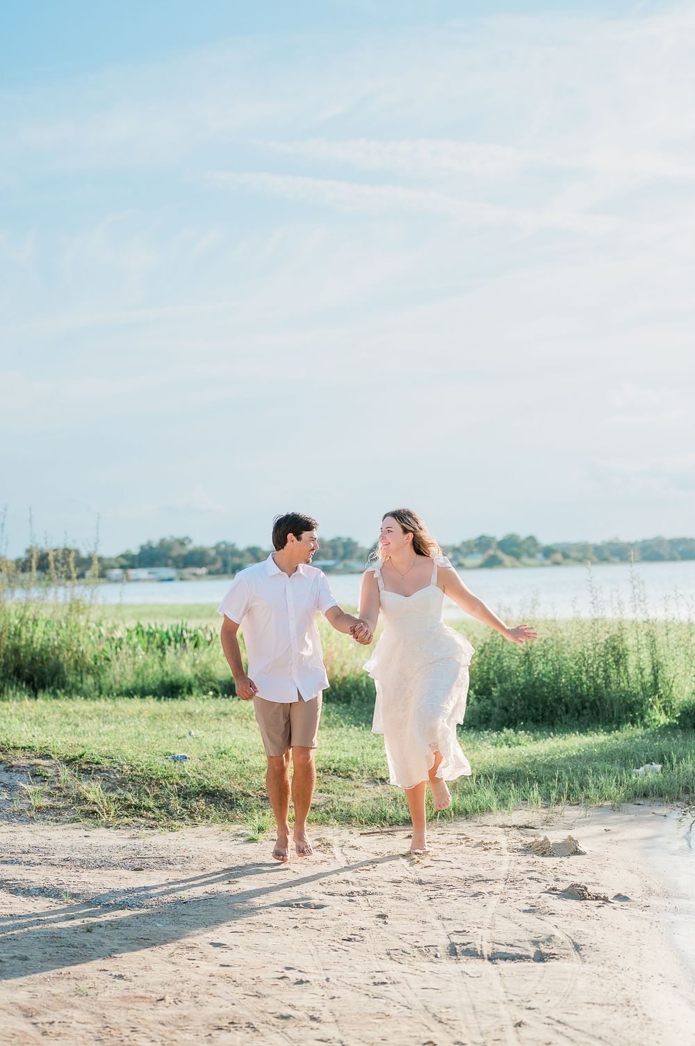 A couple in light summer outfits walks hand in hand along a sandy beach path on a bright sunny day.