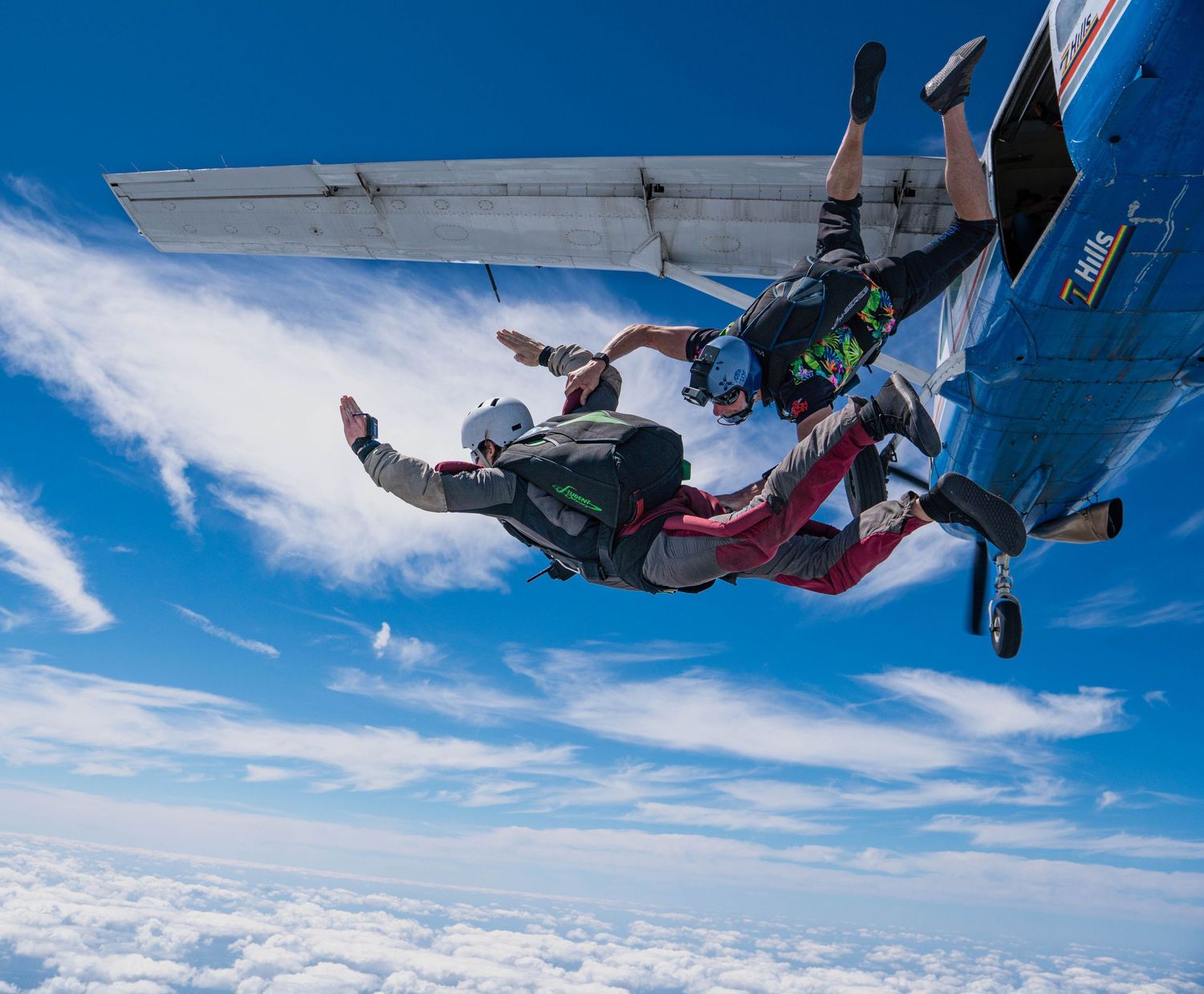 Two skydivers exit a plane at high altitude against a bright blue sky with clouds below.