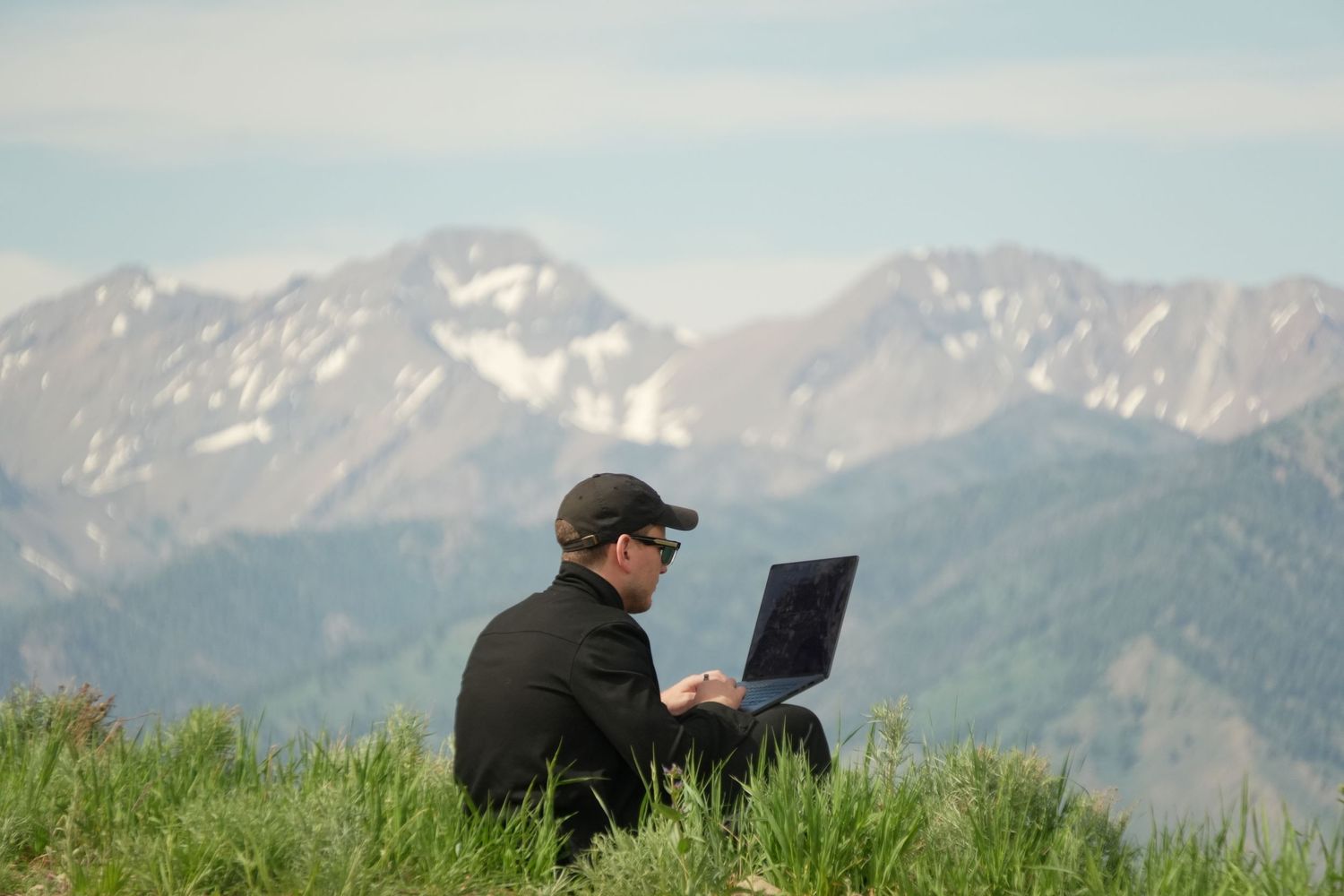 Man using laptop on a grassy hilltop with snow-capped mountain range in the background.