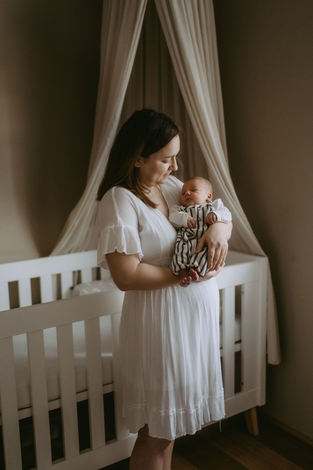 Mother and baby sharing quiet moments by a white crib with flowing curtains.