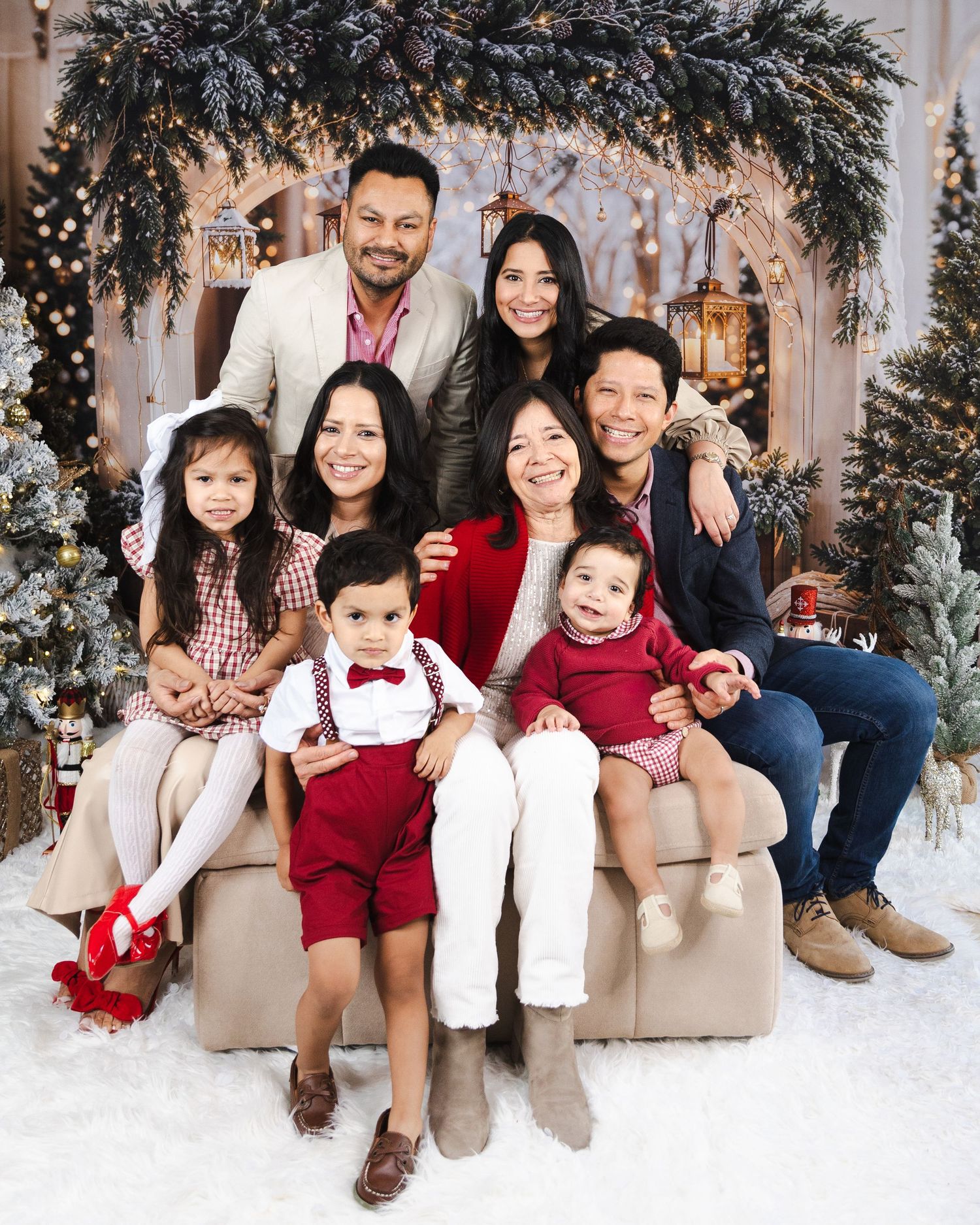 Large family group in coordinated red and neutral outfits poses together on beige couch against holiday backdrop.