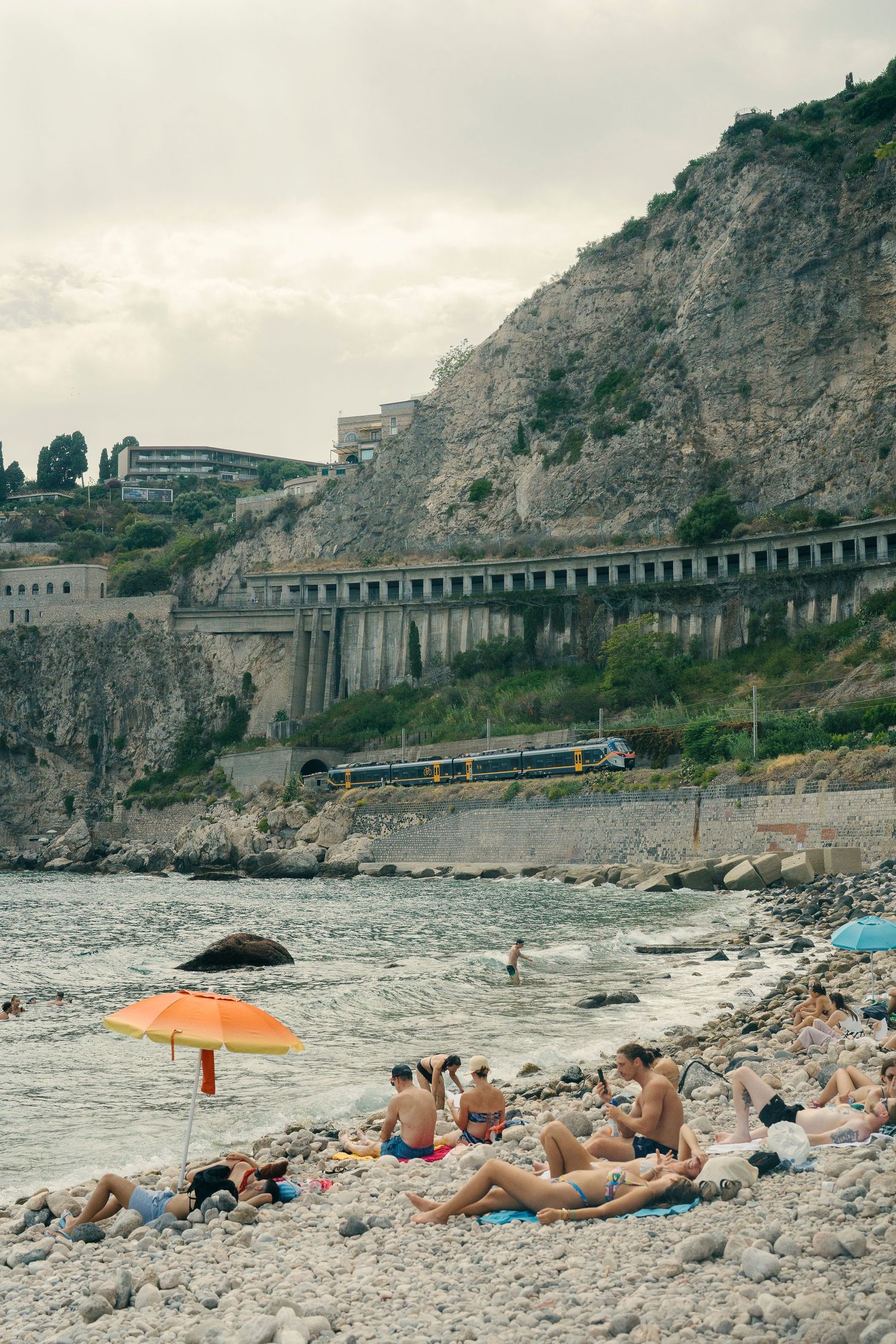 Beachgoers relax on a rocky Mediterranean coastline with steep cliffs and a railway track visible above.