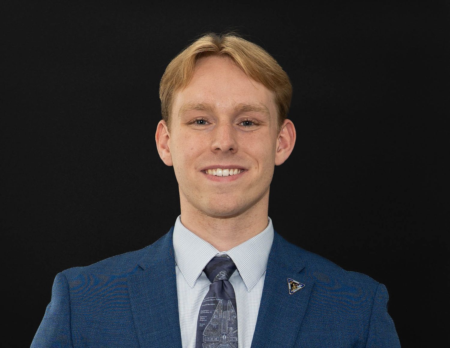 Professional portrait of someone in a blue suit with a patterned tie against a black backdrop.