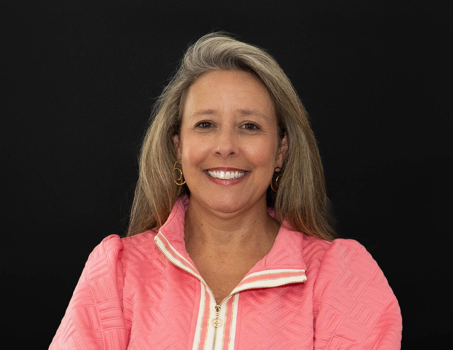 A woman in a coral pink jacket smiles warmly against a black background in a professional headshot series.