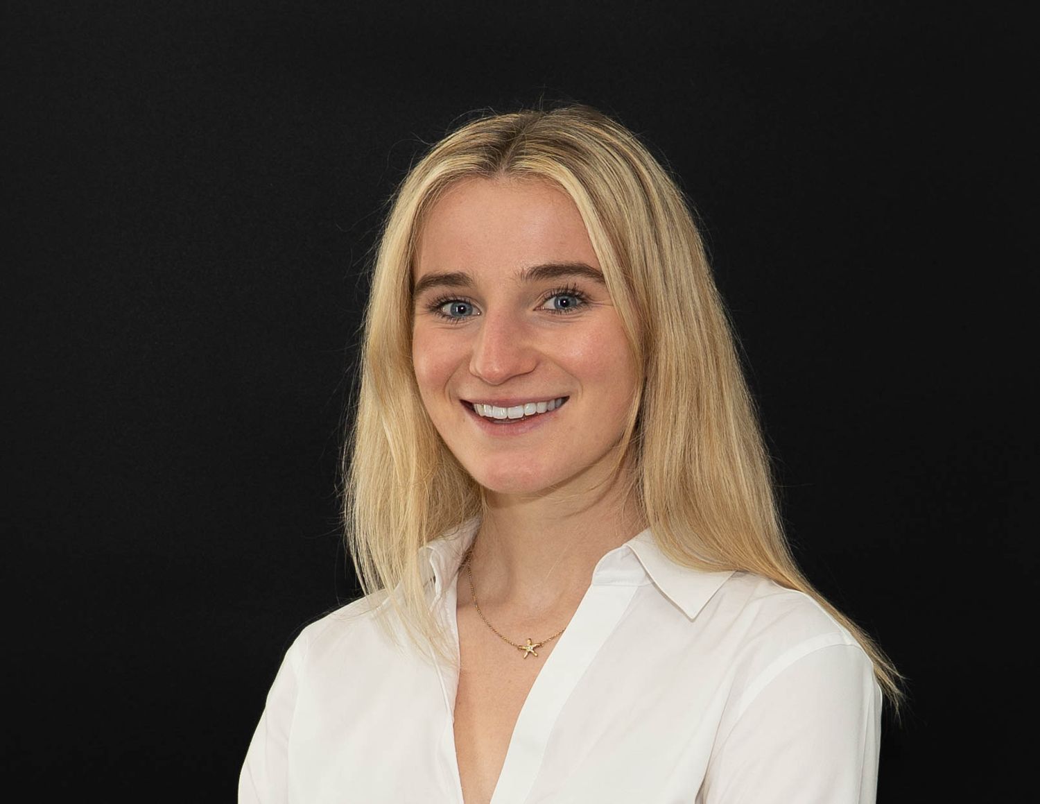 Professional headshot of a smiling person with blonde hair wearing a white button-down shirt against a dark background.