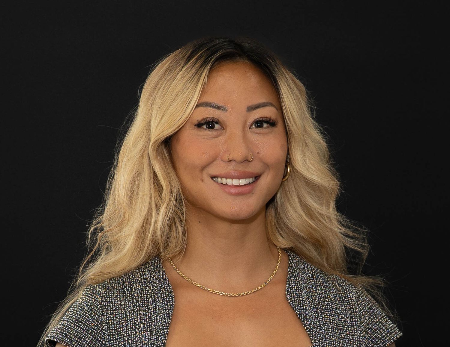 Professional headshot portrait of someone with blonde wavy hair wearing a gray cardigan and necklace against dark background.