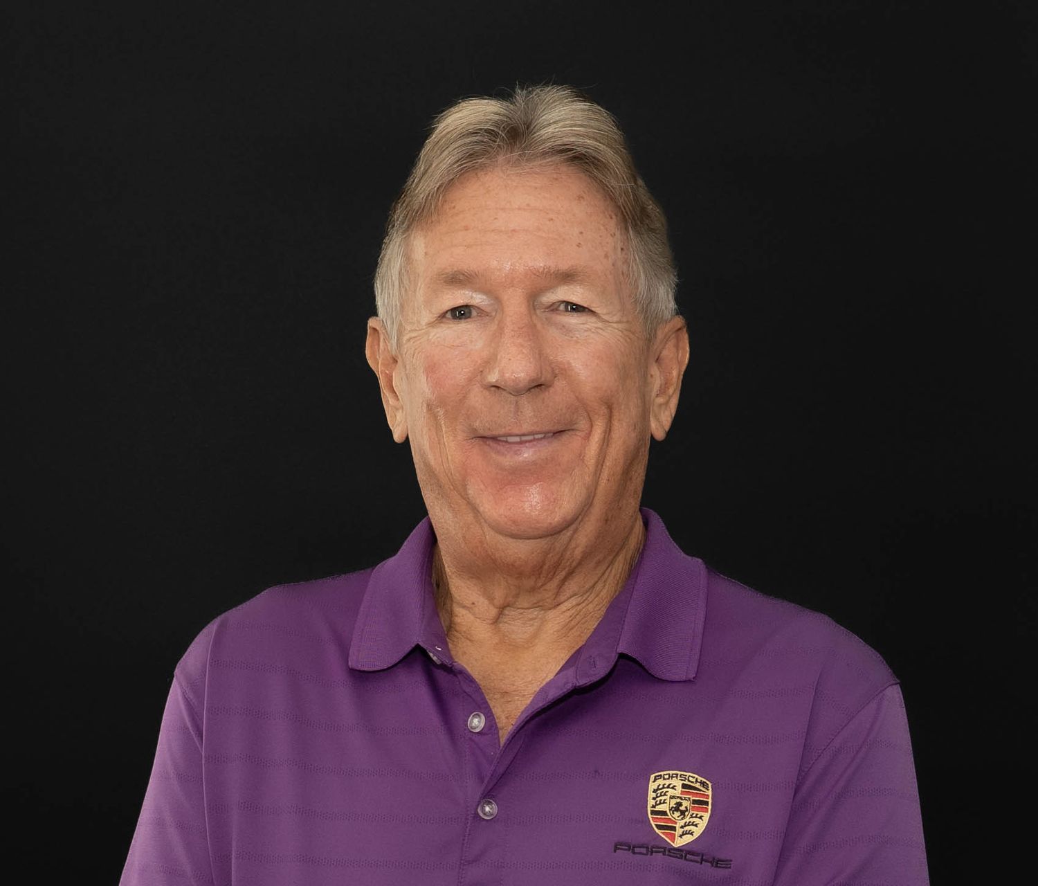 A person wearing a purple Porsche polo shirt smiles warmly against a black background.