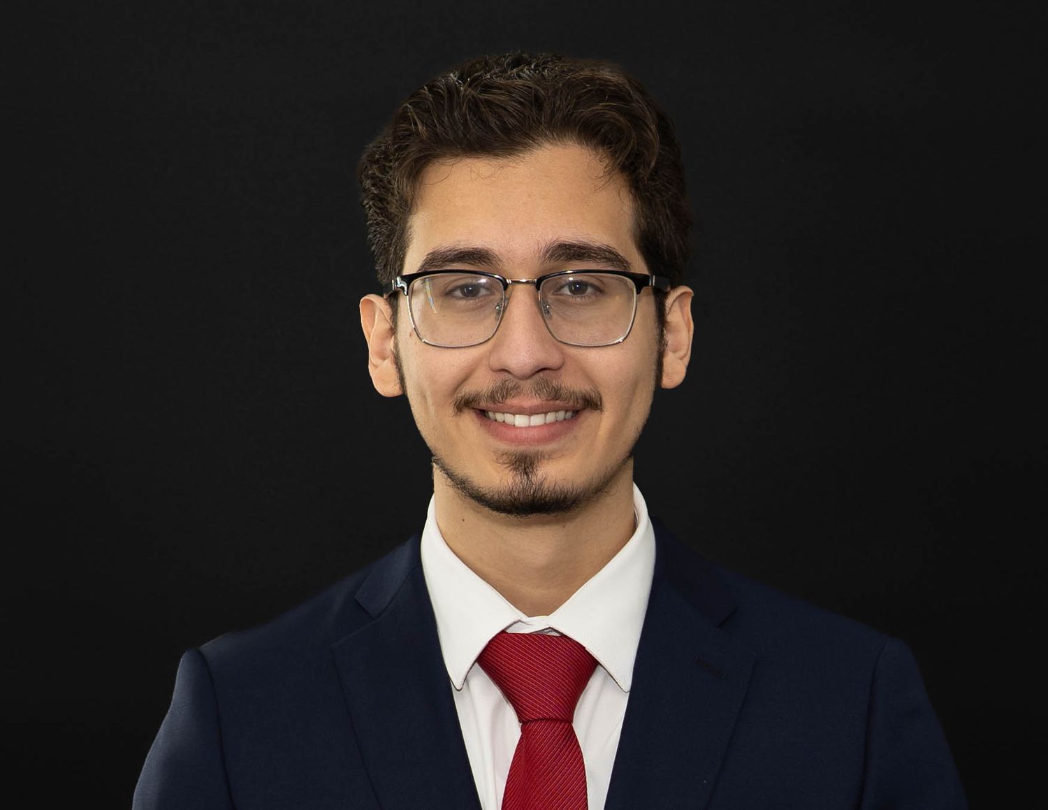 Professional headshot of someone wearing glasses, a red tie and navy suit against a dark background.