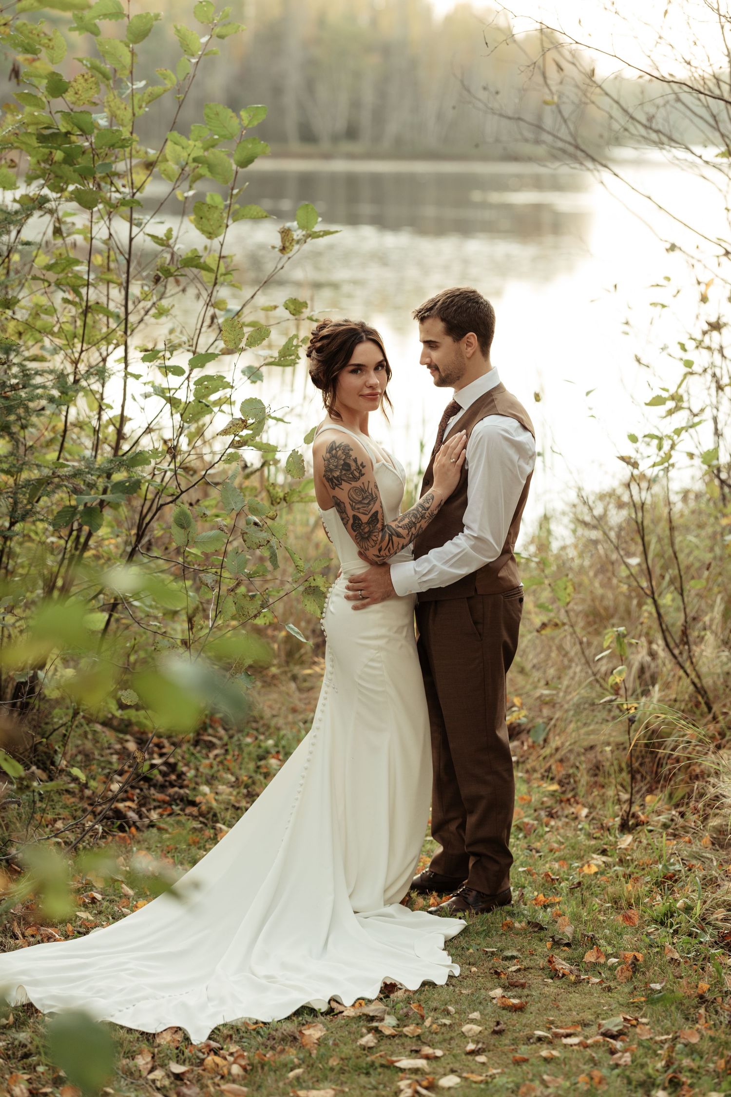 A romantic autumn wedding photo by a lake with a bride in white gown and groom in brown vest among fall foliage.