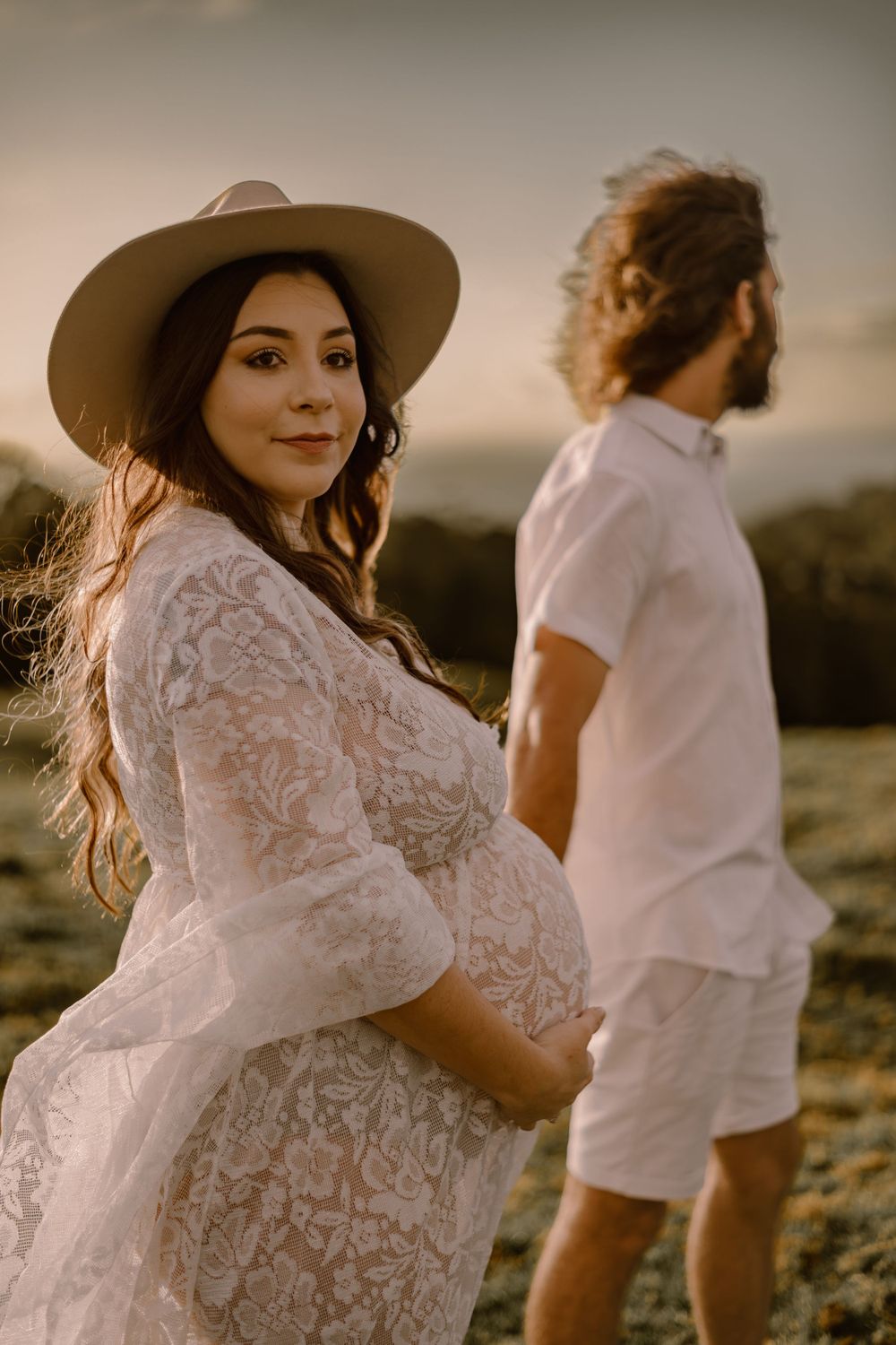 Couple in white outfits walking together on grassy hill during sunset.