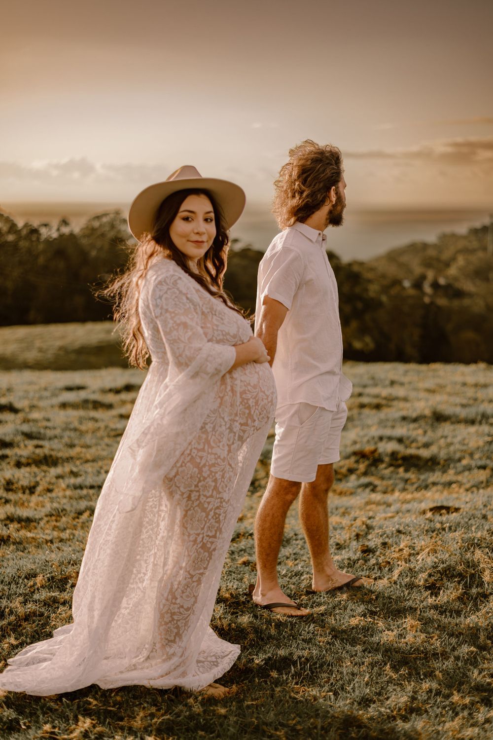 Woman in white lace dress and hat poses on grassy hillside at golden hour.