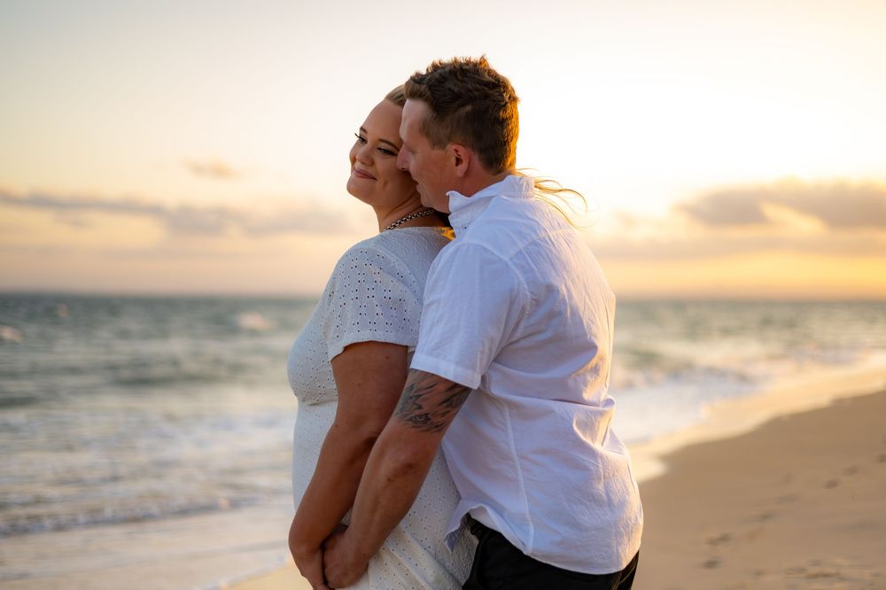 A romantic couple shares an intimate moment on the beach at sunset with ocean waves in the background.