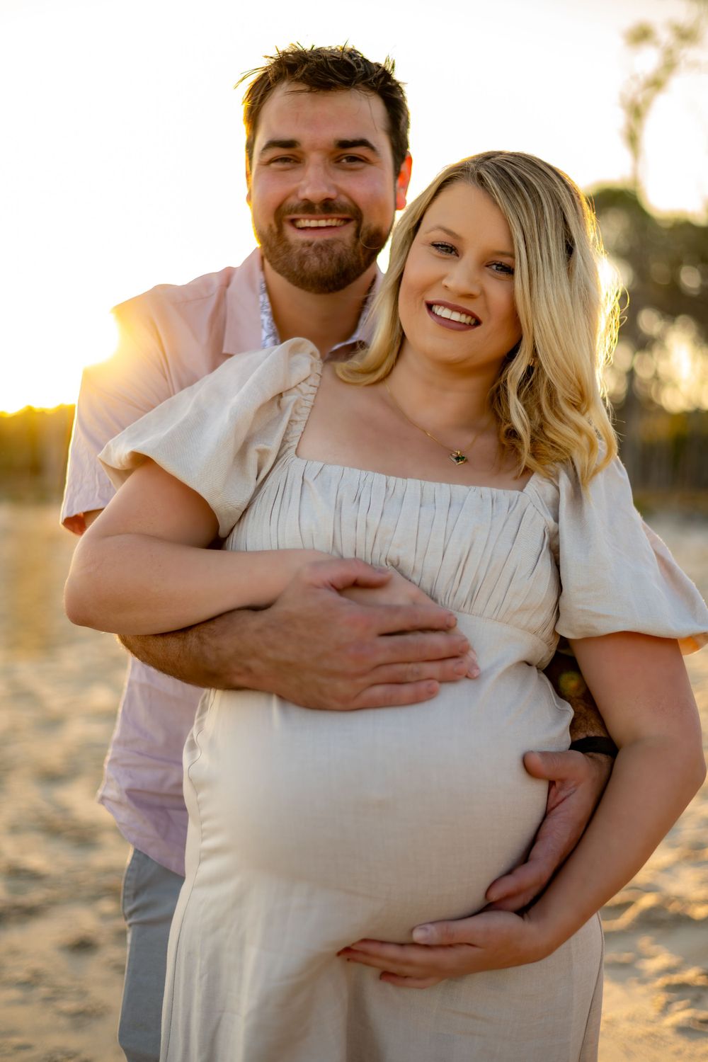 An expectant couple embraces on a sunny beach, with the woman wearing a white off-shoulder dress.