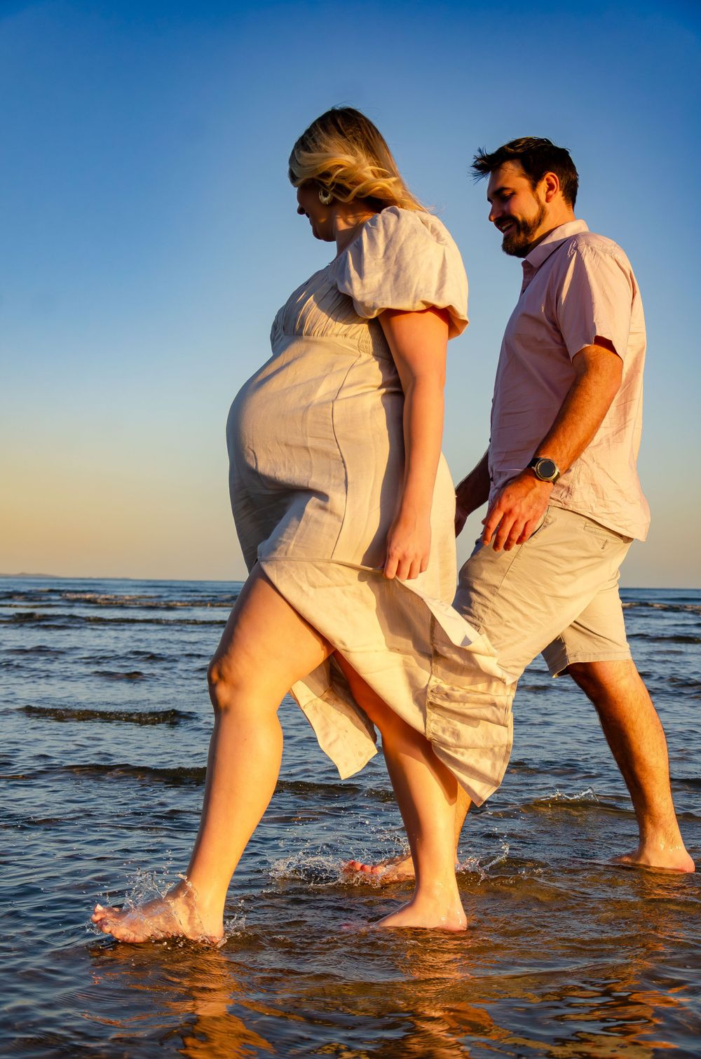 Two people in white clothing splash and play in the shallow ocean water during a beach photoshoot.