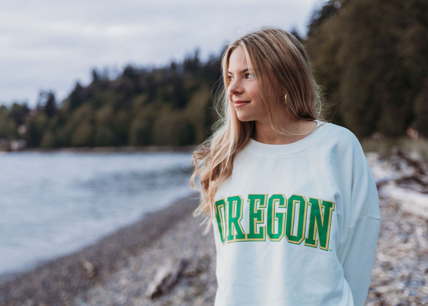 A person wearing a white Oregon sweatshirt stands near a rocky beach with evergreen trees in the background.