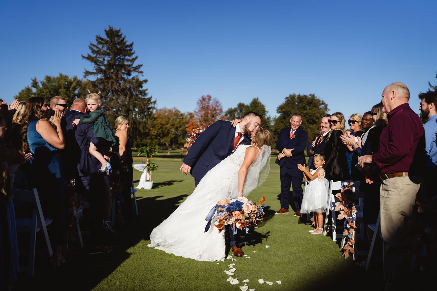 A couple shares their first kiss as newlyweds on a golf course surrounded by wedding guests on a sunny autumn day.
